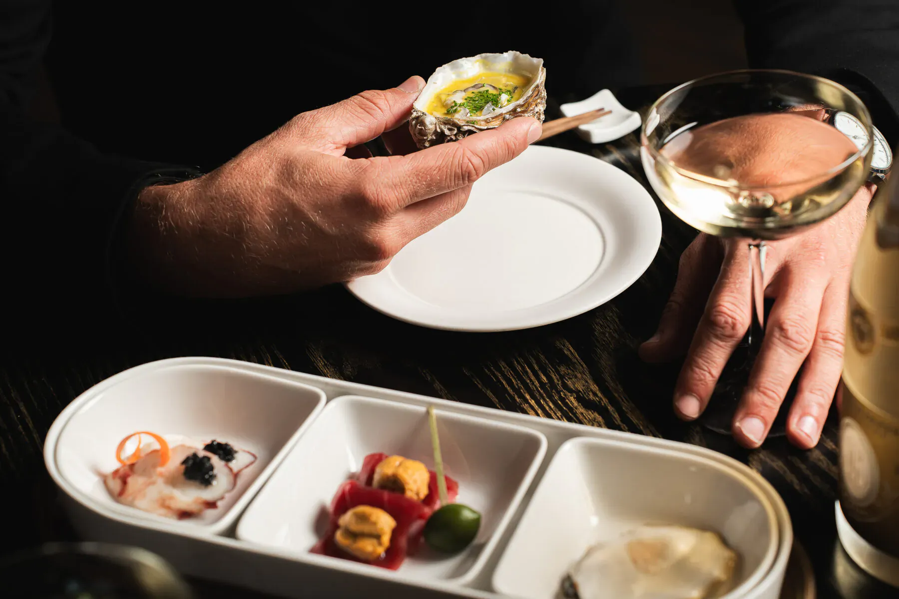 Hand holding fresh oyster on the half shell beside white wine glass and divided dish of seafood appetizers on wooden table