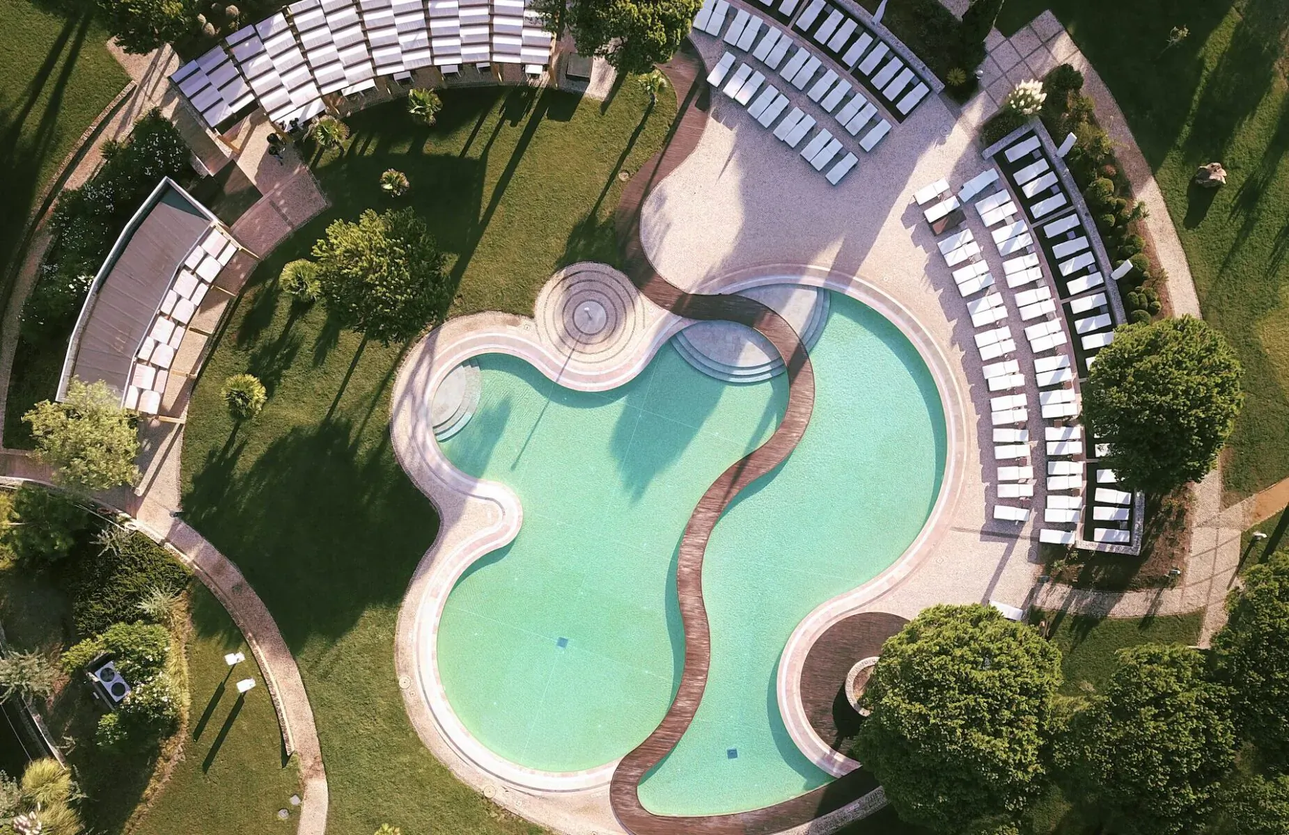 Aerial view of heart-shaped turquoise pool at Sheraton Cascais Resort, surrounded by white lounge chairs and lush green lawns.