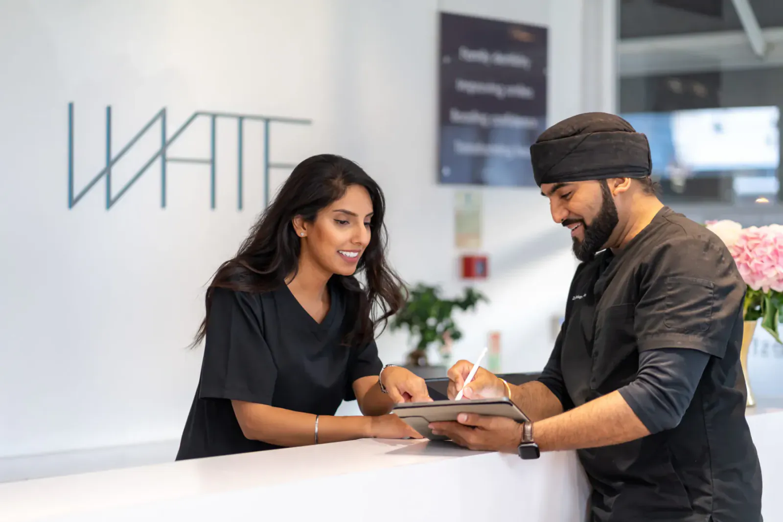 At White Dental reception, female staff in black uniform discusses tablet with male colleague in black uniform and turban.