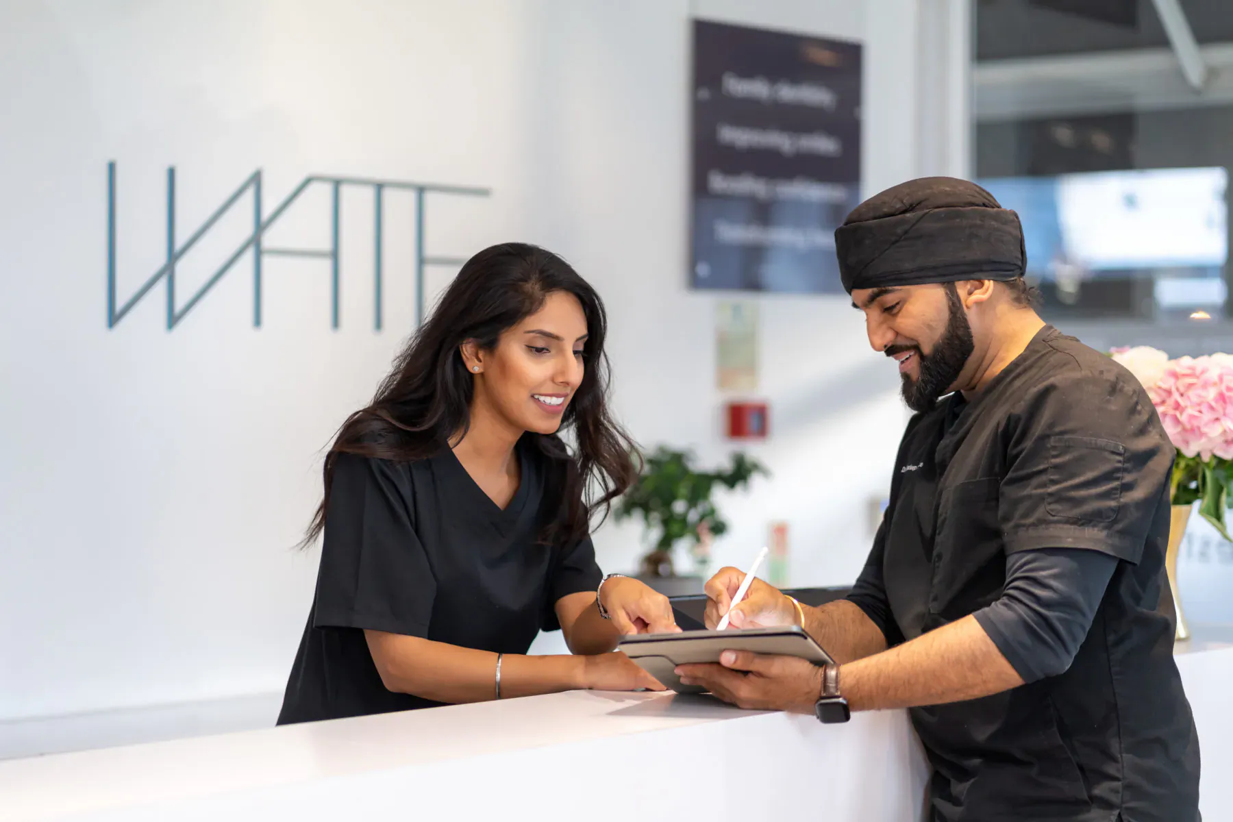 At White Dental reception, female staff in black uniform discusses tablet with male colleague in black uniform and turban.