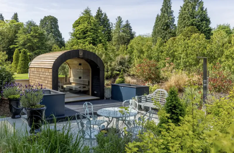 Wooden arched pavilion with white benches and table in lush garden patio at PoB Hotels.
