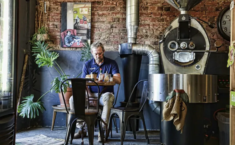 Man in blue shirt sits at table with cup in rustic Kalk Bay cafe beside large coffee roaster and plants