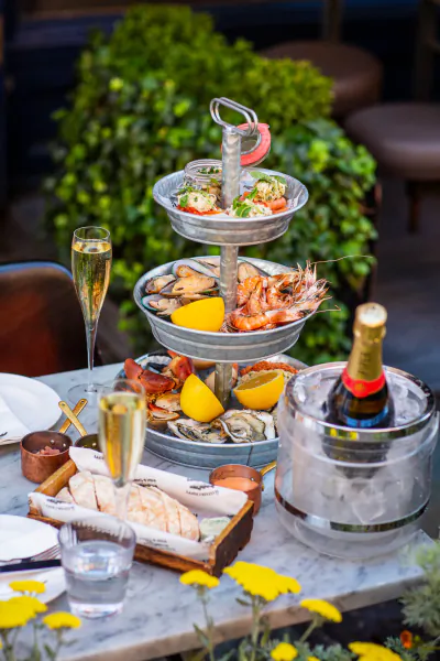 Three-tier seafood tower with oysters, prawns, lemon on marble table in Lobster Pot Booth, with champagne bottle in ice bucket and glass beside potted plants.