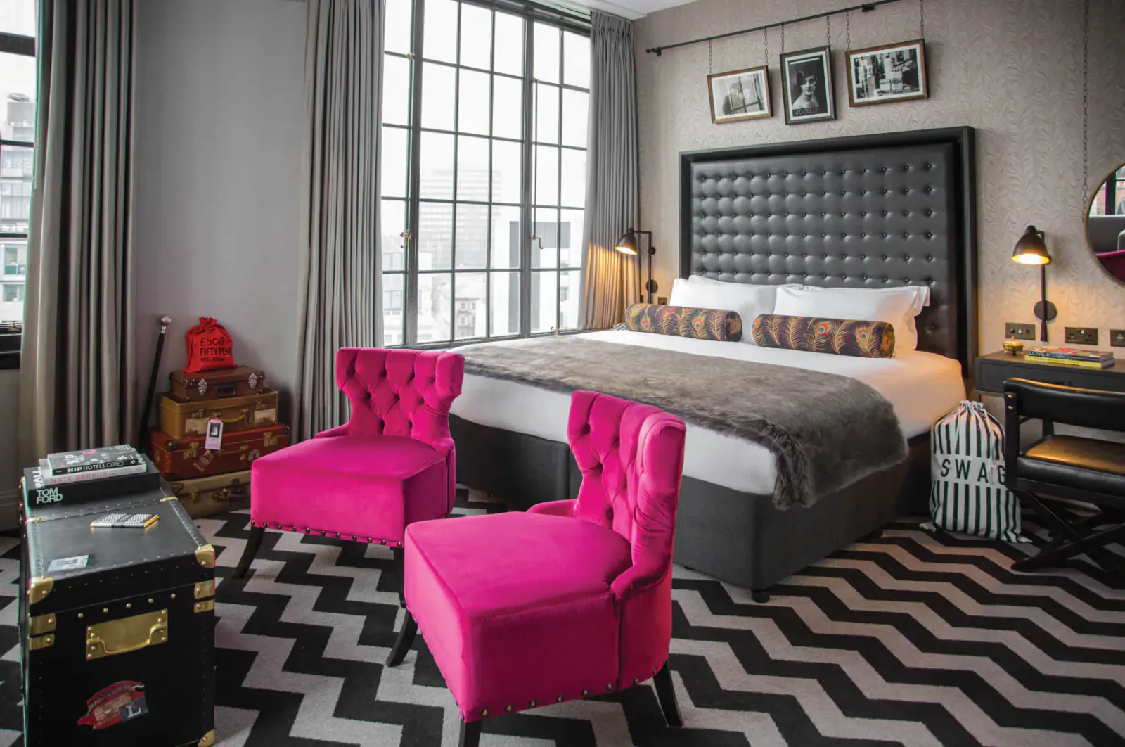 Plush Hotel Gotham bedroom with gray tufted bed, pink velvet chairs, vintage suitcases, black zig-zag rug, large window.
