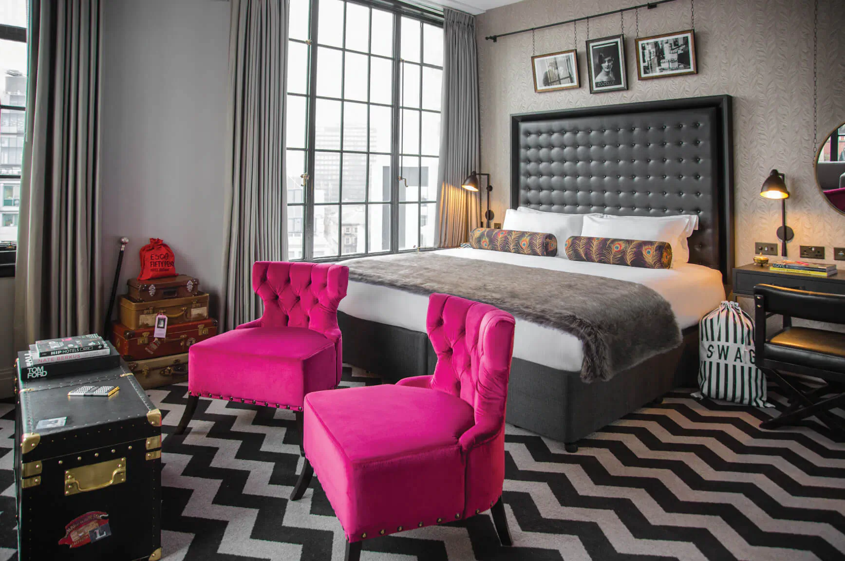 Plush Hotel Gotham bedroom with gray tufted bed, pink velvet chairs, vintage suitcases, black zig-zag rug, large window.
