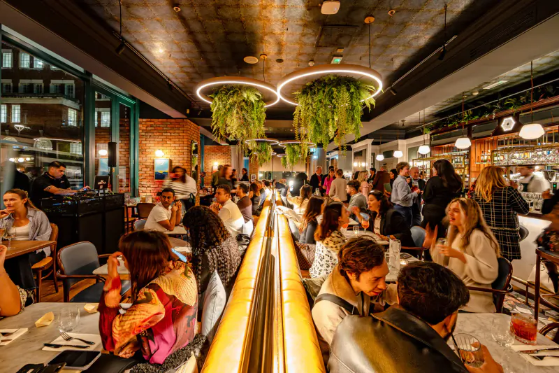 Crowded Amalfi Ristorante dining room with diverse patrons at long sushi conveyor belt table under hanging plants and neon lights