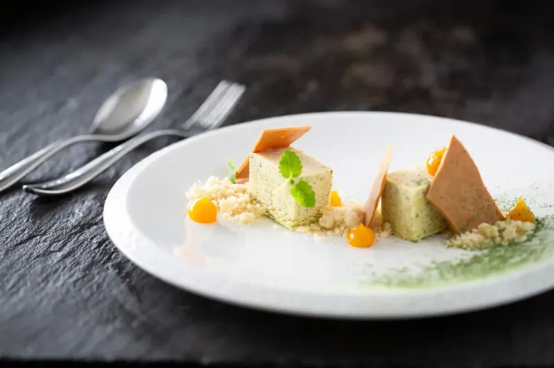 Elegant plated dessert with white tofu cubes, orange kumquats, herbs, and crisps on black slate with silverware