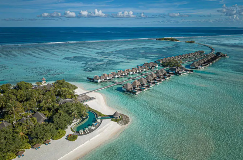 Aerial view of overwater bungalows and infinity pool on a lush Maldives resort island with turquoise lagoon and ocean.