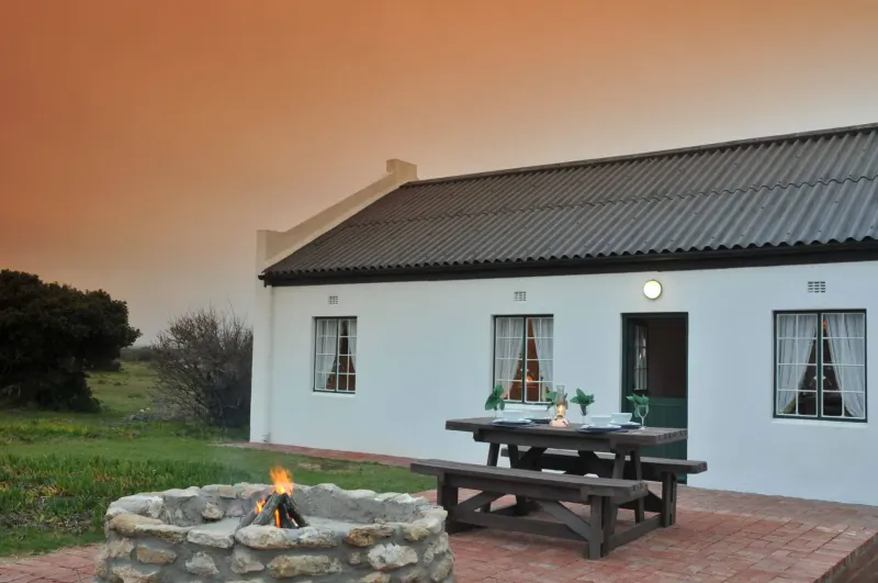 White cottage with gabled roof at De Hoop Preserve, picnic table by fire pit in yard at sunset.