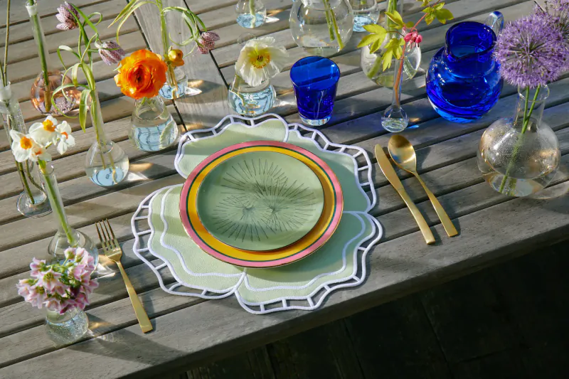 Luxury table setting on wooden surface with green Truffle Bee plate, gold flatware, blue glasses, and colorful flowers in vases.