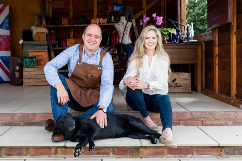 Smiling man in leather apron and blonde woman with black Labrador dog outside wooden shop with bags, Union Jack flag, and crates.