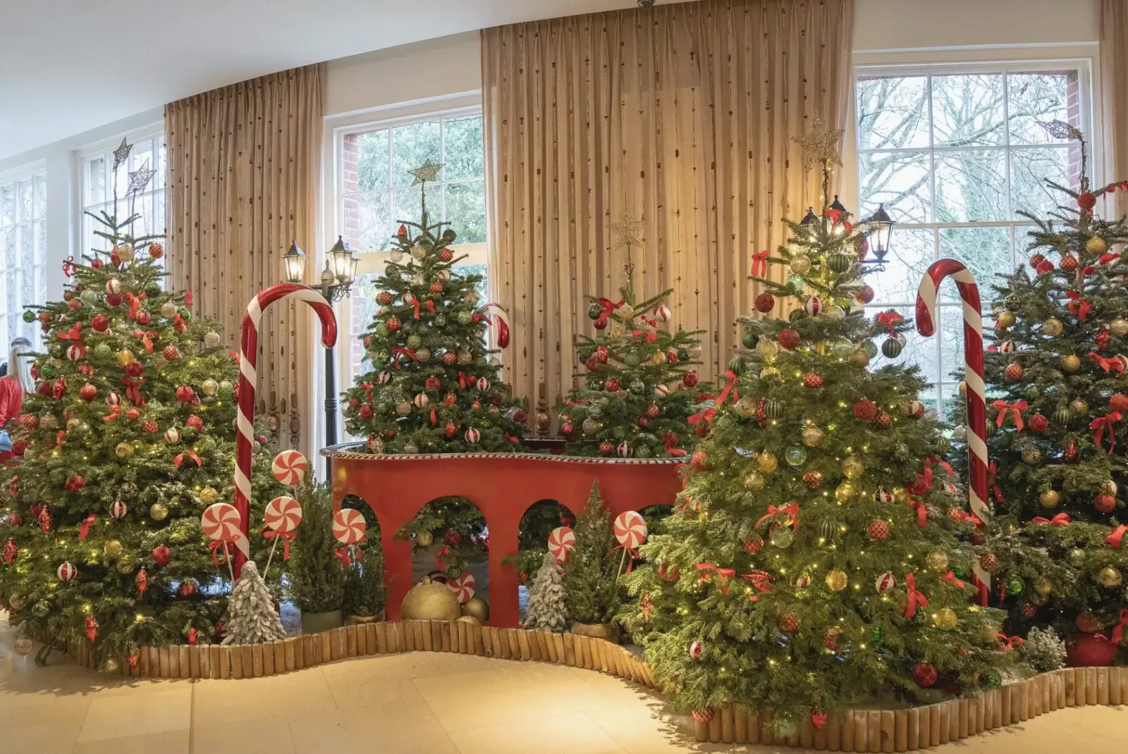 Festive indoor Christmas display with decorated trees, giant candy canes, lamppost, and arched red platform amid large windows.