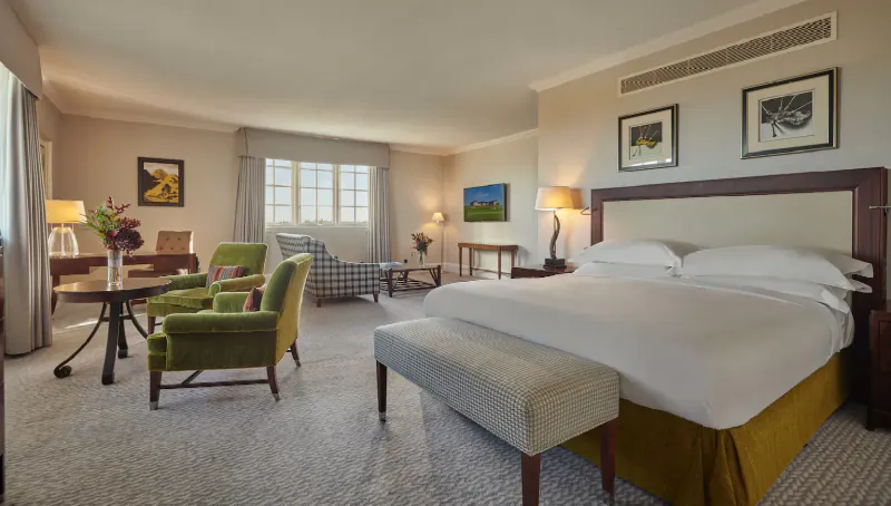 Luxurious hotel suite with king bed, green armchairs, desk, bench, and landscape artwork on walls.