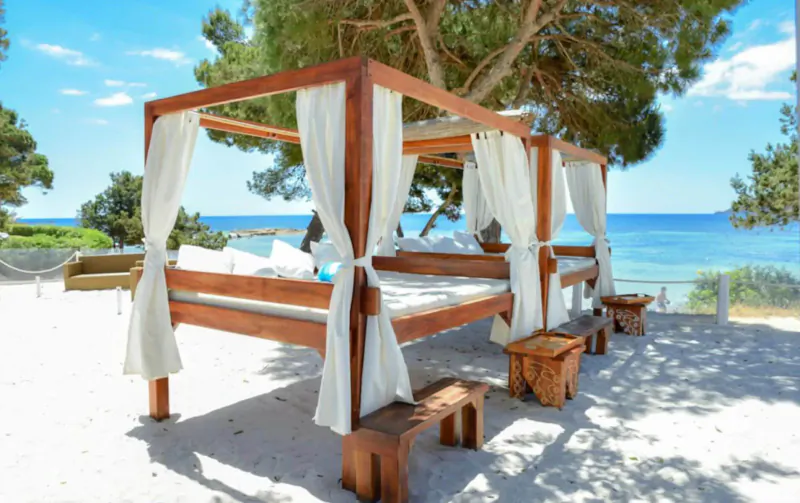 Wooden beach bed with white curtains on sandy shore at Nikki Beach Ibiza, pine trees, sea view.