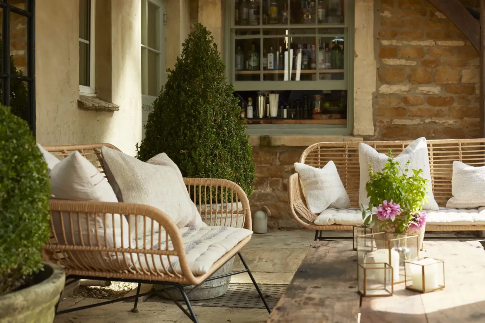 Cozy outdoor patio with rattan chairs, white cushions, potted conifers, flowers, and candles beside stone cottage window.
