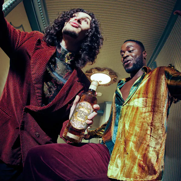 Two stylish men posing dynamically with limited-edition Havana Club x Jonathan Raksha bottle in gallery setting