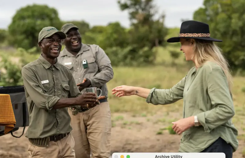 Two Black male rangers in khaki uniforms and a blonde woman in green blouse exchange cups outdoors in Timbavati bushveld.