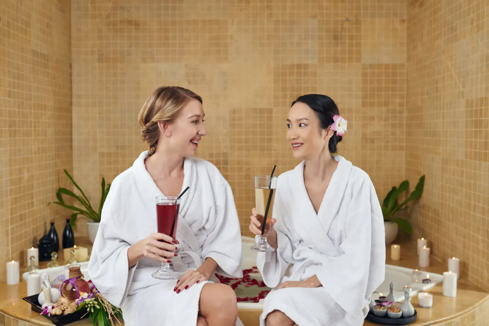 Two smiling women in white robes clink purple and orange drinks in a luxurious spa hot tub with candles and flowers.