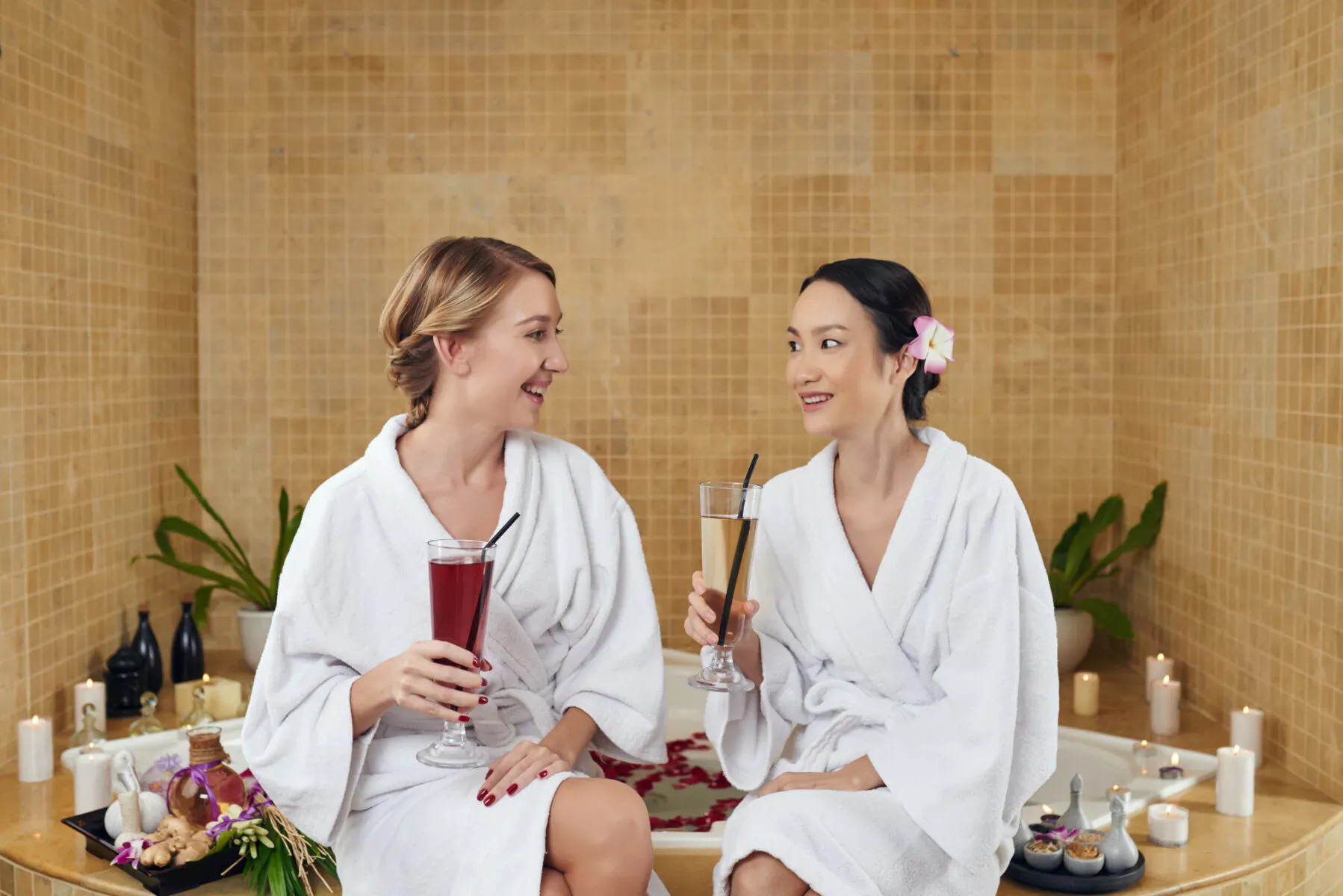 Two smiling women in white robes clink purple and orange drinks in a luxurious spa hot tub with candles and flowers.
