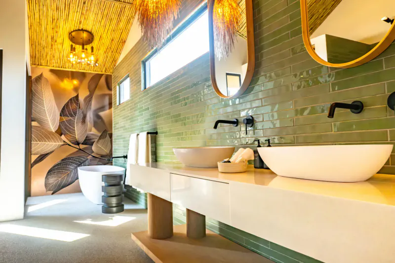 Modern bathroom with green leafy wall mural, double white vessel sinks on wooden vanity, black faucets, and thatched ceiling