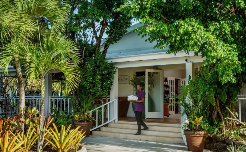Staff member holding folder walks up steps to entrance of Spice Island Beach Resort, surrounded by lush tropical plants and palms.