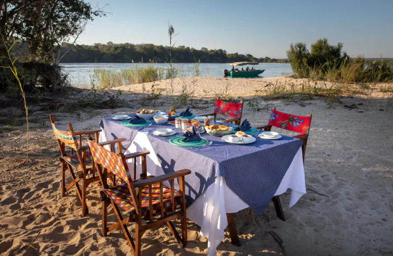 Elegant dining table set on sandy riverbank with Zambezi River and green boat in sunset backdrop, Livingstone Zambia