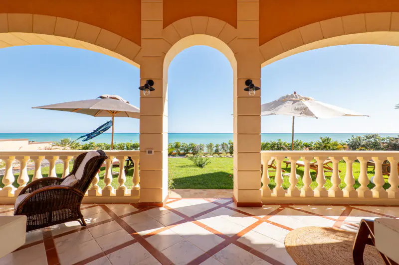 Luxurious terracotta arched terrace with wicker armchair, umbrellas, lush garden, and sunny Sicily sea view.