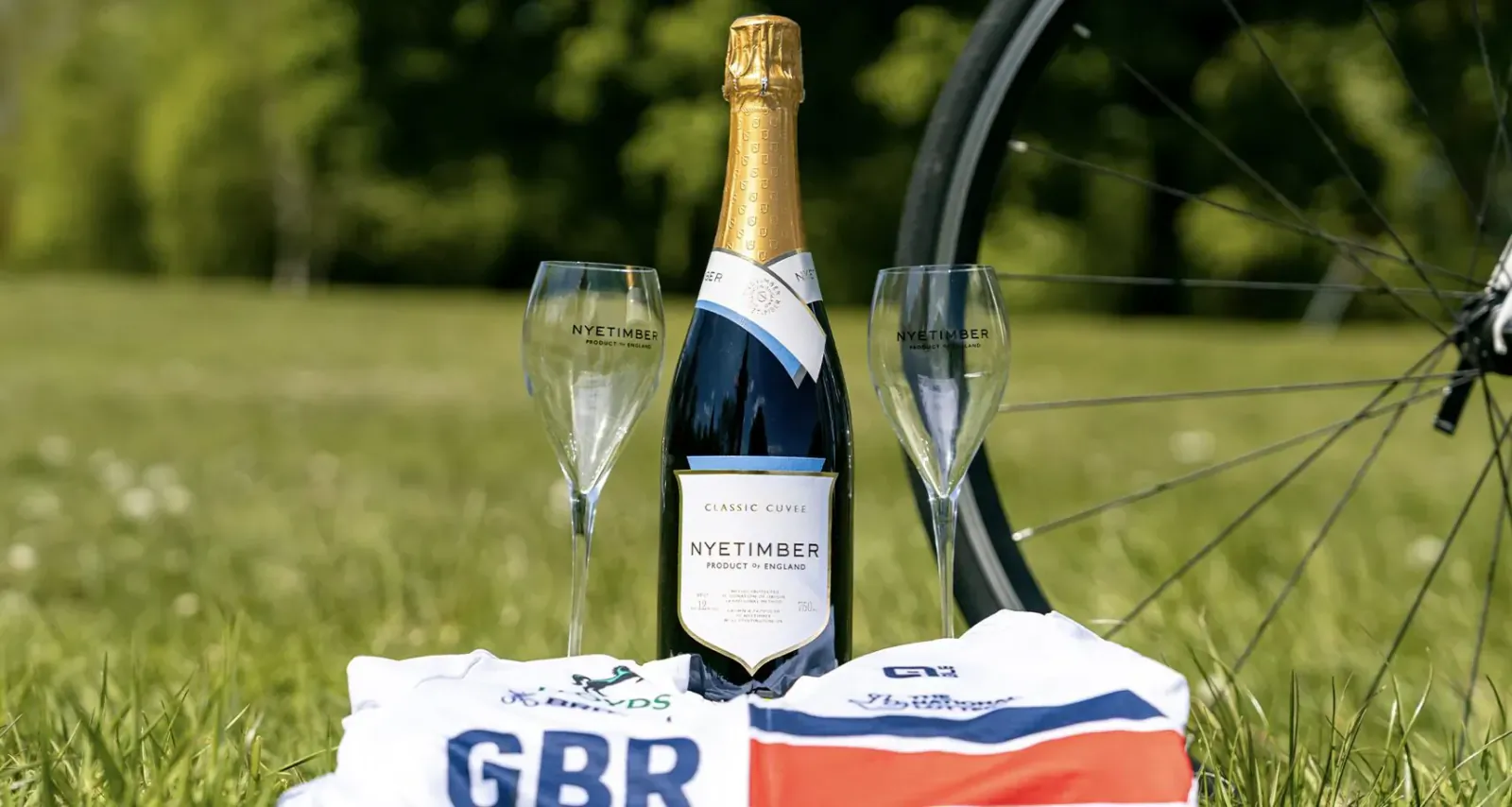 Nyetimber sparkling wine bottle with two glasses beside bicycle wheel, GBR jersey, and box on green grass.