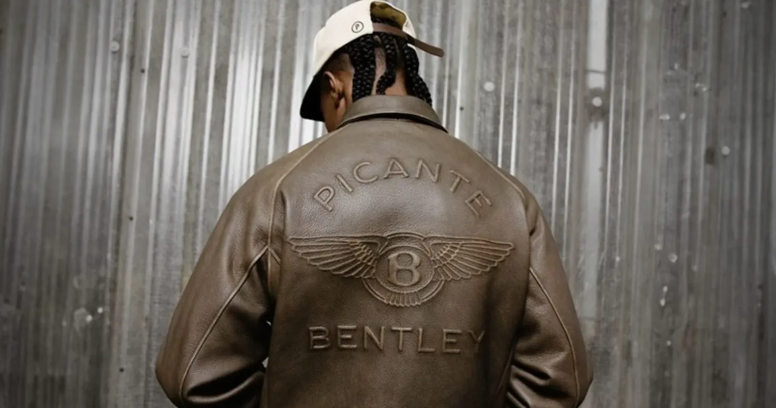 Man in brown Bentley Picante leather jacket with '8' emblem, white cap with dreads, standing against corrugated metal wall