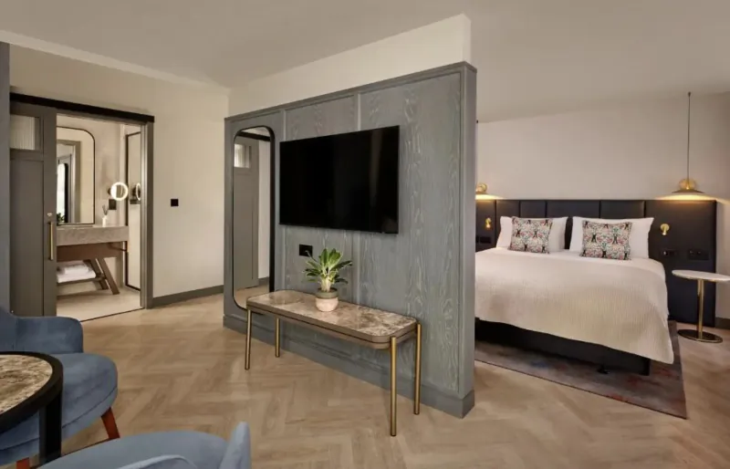 Modern hotel room at The Reach at Piccadilly, Manchester, with king bed, TV on gray partition, en-suite bathroom, and velvet chairs.
