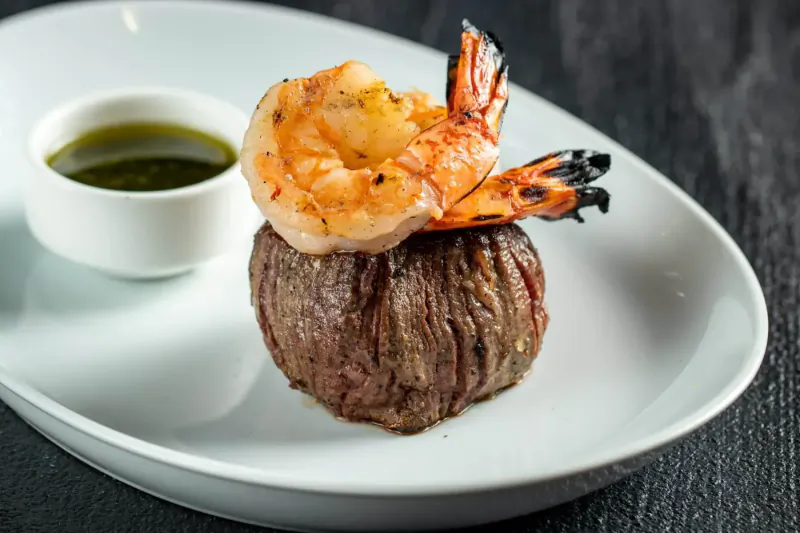Grilled filet mignon topped with seared shrimp and side of green sauce on white plate
