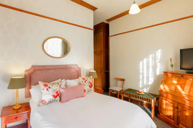 Cozy hotel bedroom with pink upholstered bed, floral pillows, wooden furniture, oval mirror, and TV.