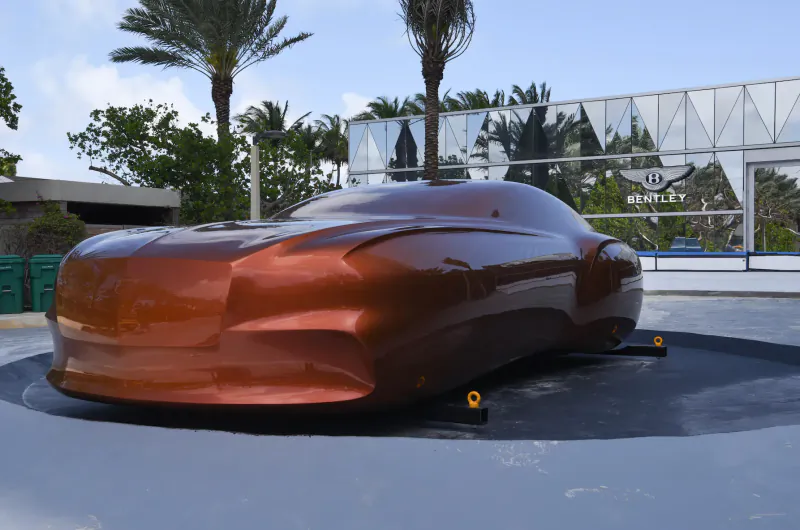 Shiny bronze Bentley concept car on rotating platform at Bentley Residences gallery, palm trees and glass building backdrop.