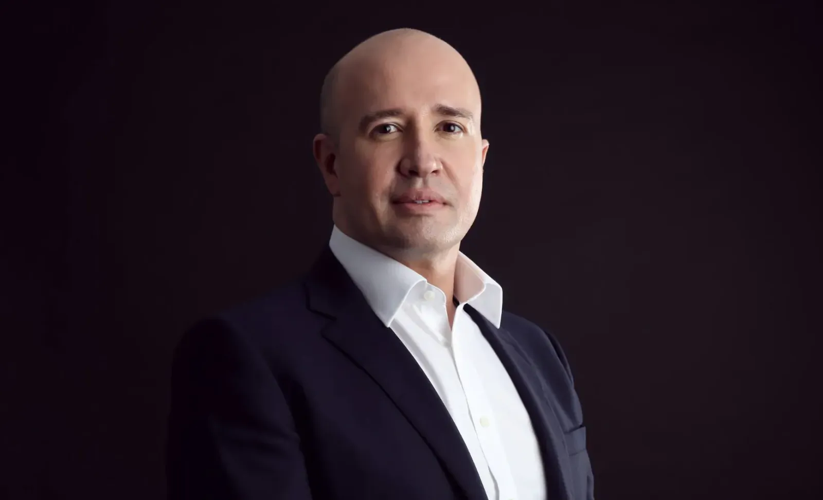 Dr. Joney de Souza, bald man in navy suit and white shirt, professional headshot on dark background