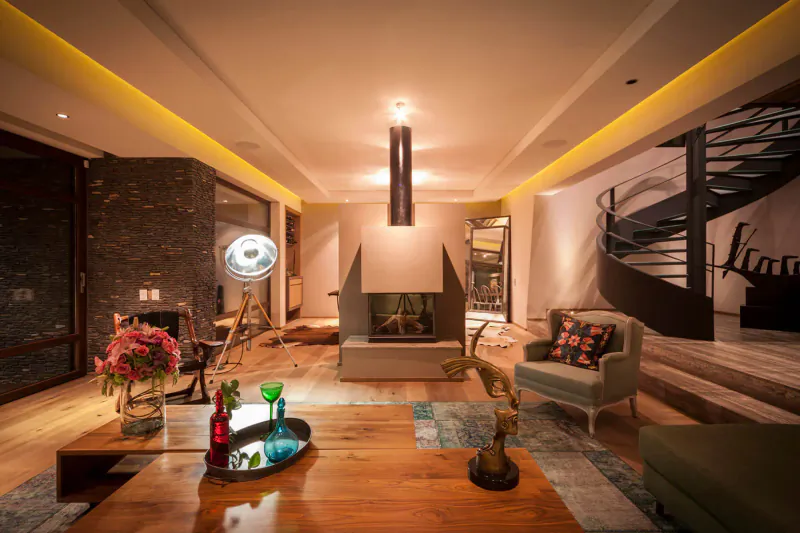 Modern living room with central wood table, colorful flowers and bottles, double-sided fireplace, fan, armchair, and spiral staircase