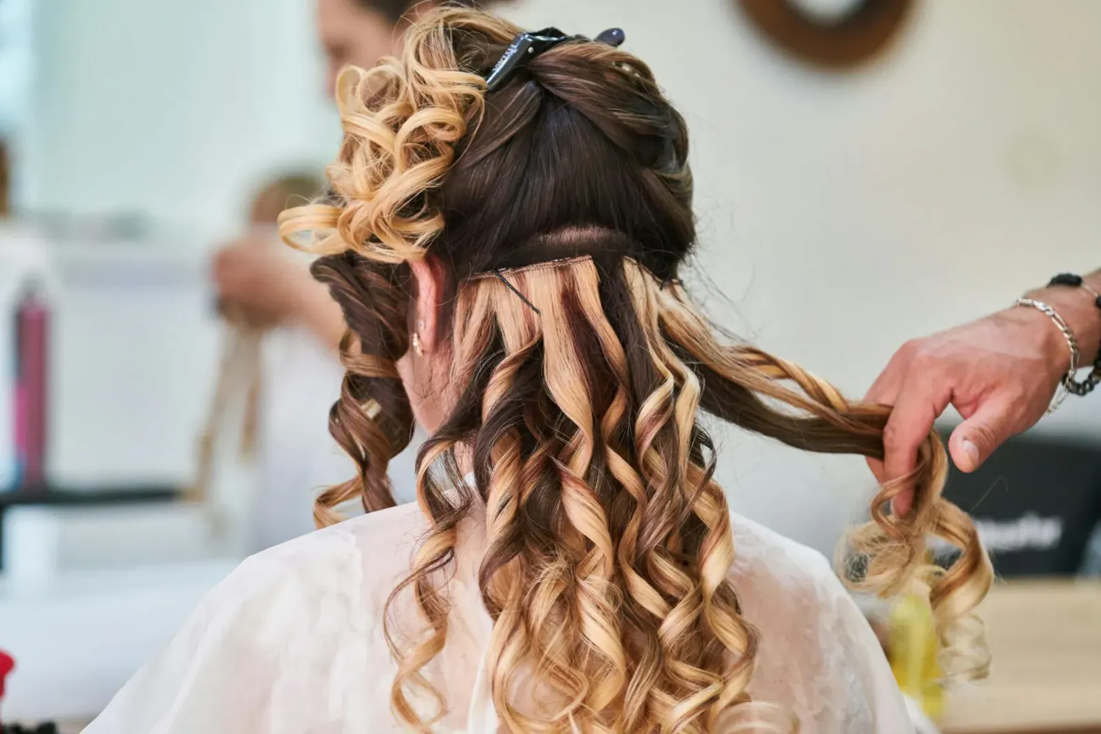 Stylist braiding blonde and brown hair weave into woman's long wavy hairstyle in salon mirror reflection