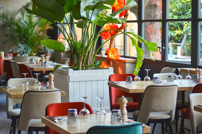 Elegant dining area at Lou Calen with wooden tables, colorful chairs, wine glasses, lush green plants, and large arched windows