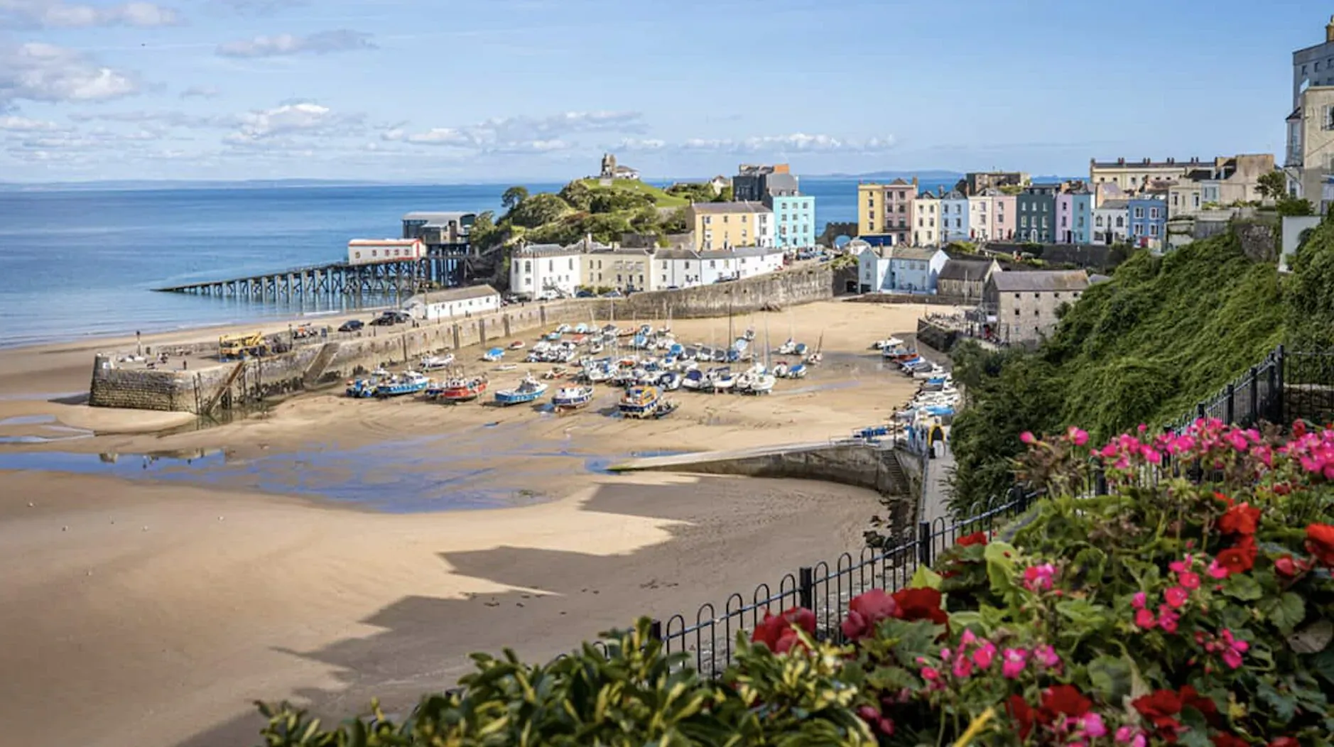 Panoramic view of Tenby's colorful harbor, sandy beach, pier, boats, and sea under blue skies