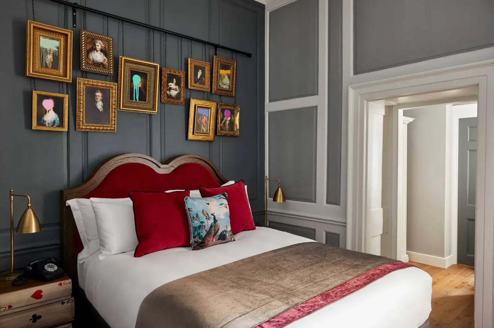 Luxurious hotel bedroom with red tufted headboard, white bedding, gold-framed portraits on gray wall, and brass lamps