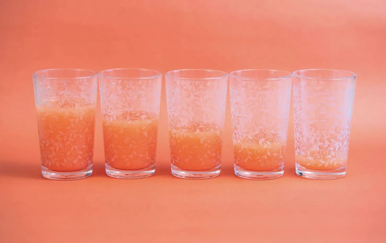 Five clear glasses with orange juice at varying fill levels on pink background, for juice cleanse.