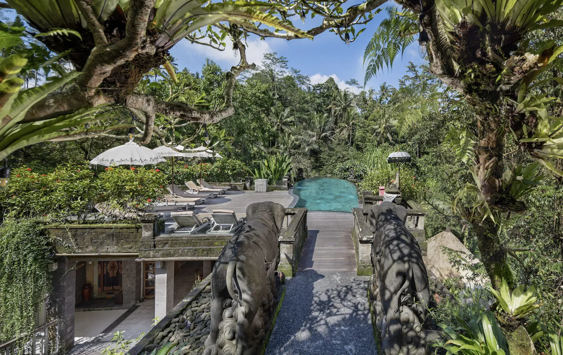 Kayon Resort Bali: Stone bridge over turquoise pool, framed by lush tropical greenery and Balinese umbrellas