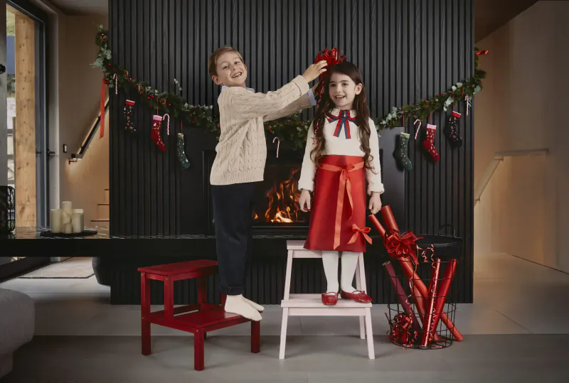 Boy stands on red stool placing red bow in girl's hair by modern fireplace with Christmas garlands and stockings.