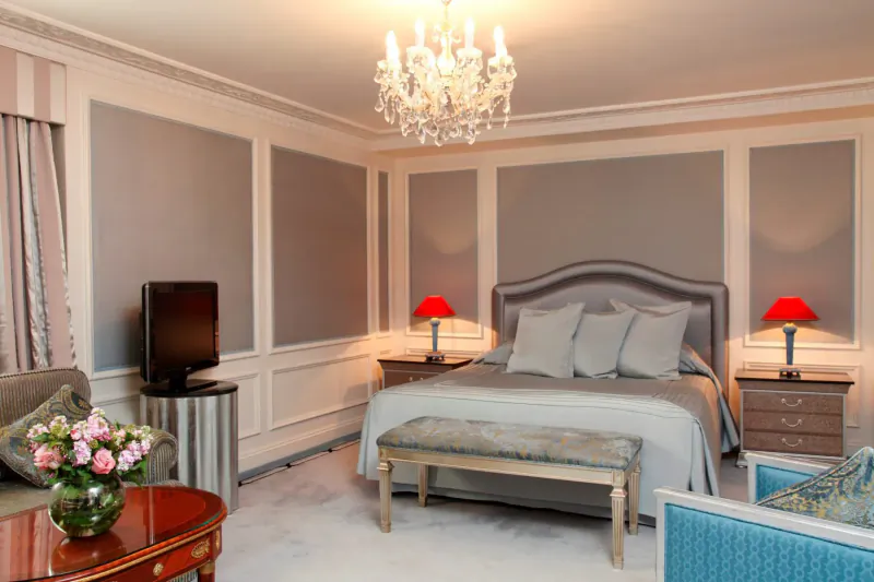 Elegant Bentley Hotel bedroom with chandelier, gray upholstered bed, red lamps, TV, flowers, and armchairs.