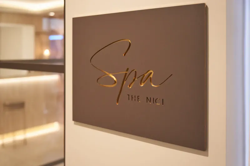 Elegant wall-mounted sign reading 'Spa The NICI' in gold cursive on brown panel in modern hotel lobby.