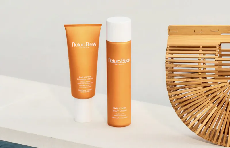 Natura Bissé orange sun protection tube and bottle with 'UVA/UVB' text beside wicker bag on white surface.