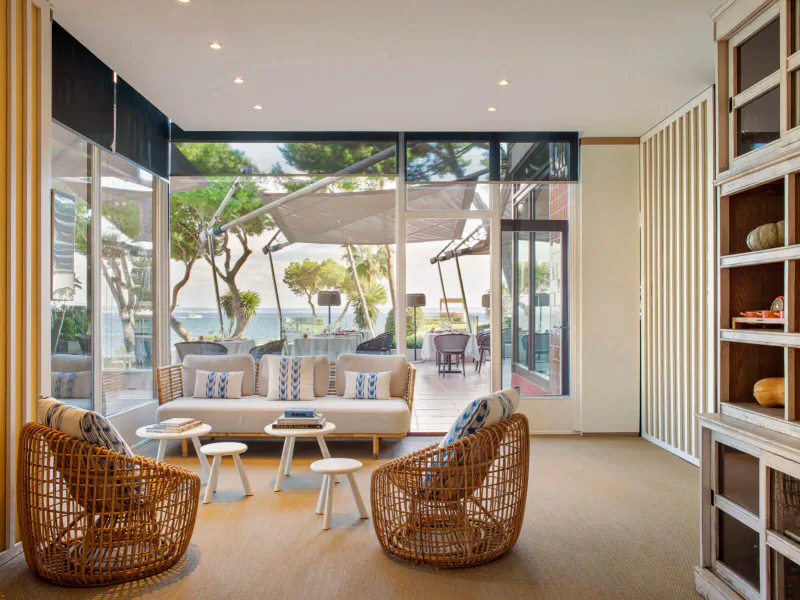 Bright indoor lounge with wicker chairs, low tables, and large windows overlooking palm trees, sea, and terrace at ARRELS by Marga Coll.