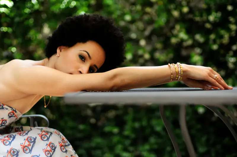 Woman with curly afro leaning on table outdoors, showcasing gold bracelets on arm, floral dress, green background