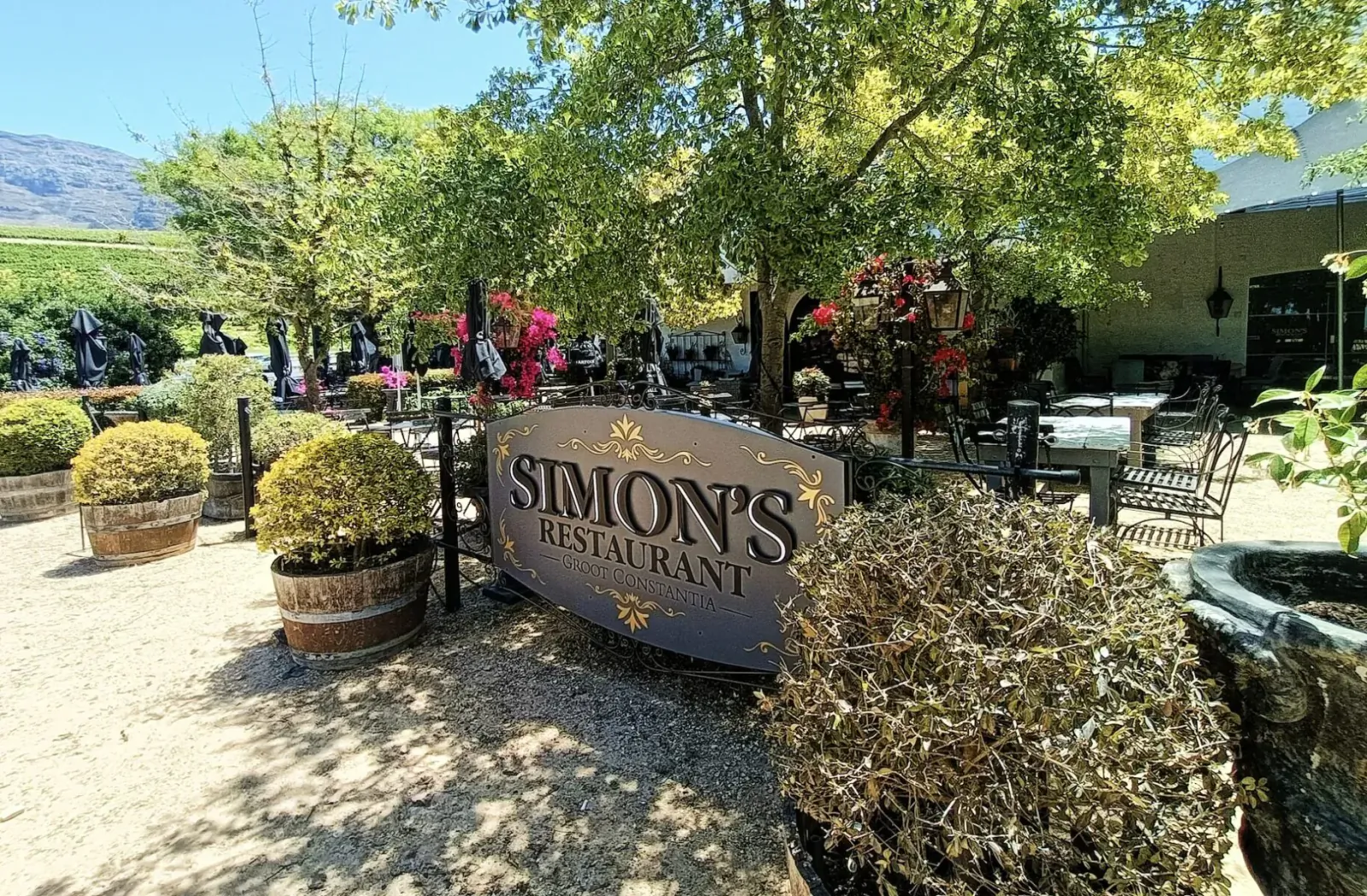 Simon's Restaurant sign in sunny outdoor patio with trees, mountains, plants, tables, and patrons.