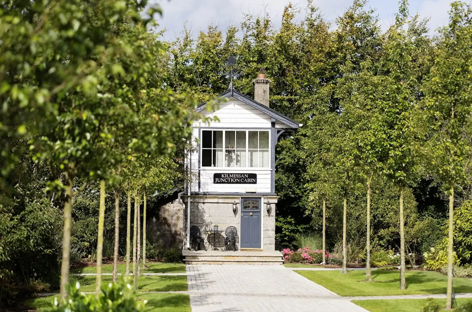 White signal box with chimney and sign, framed by tall trees and garden path in Boyne Valley, Ireland