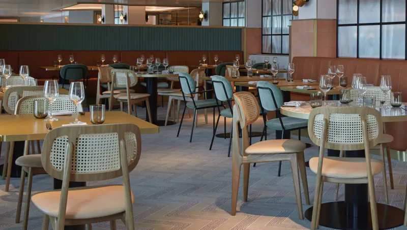 Modern adults-only cruise dining area with wooden tables, rattan chairs, glassware, and ambient lighting.